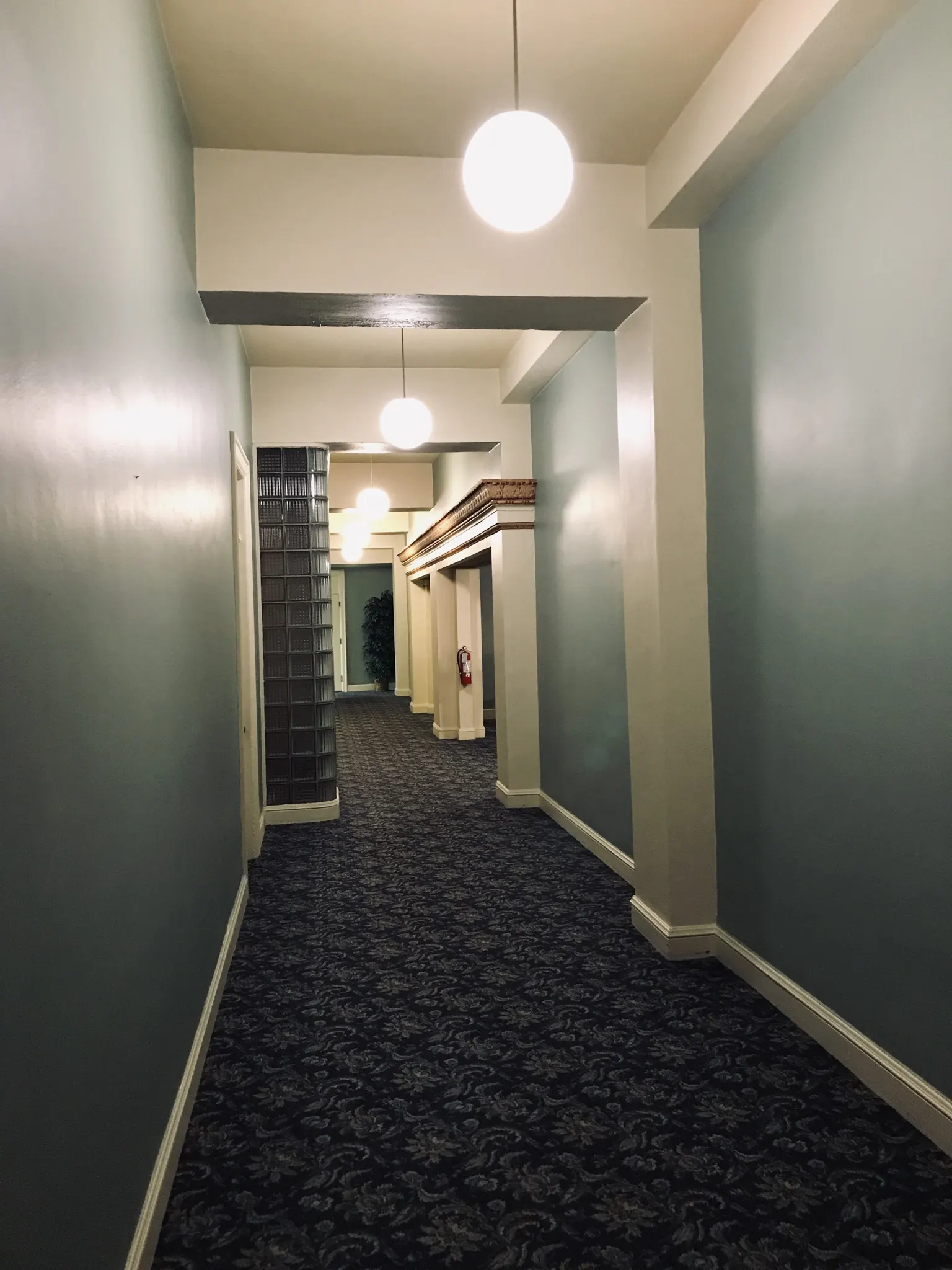 Empty hotel corridor with dark patterned carpet, pale green walls, globe pendant lights, and doorways receding into the distance