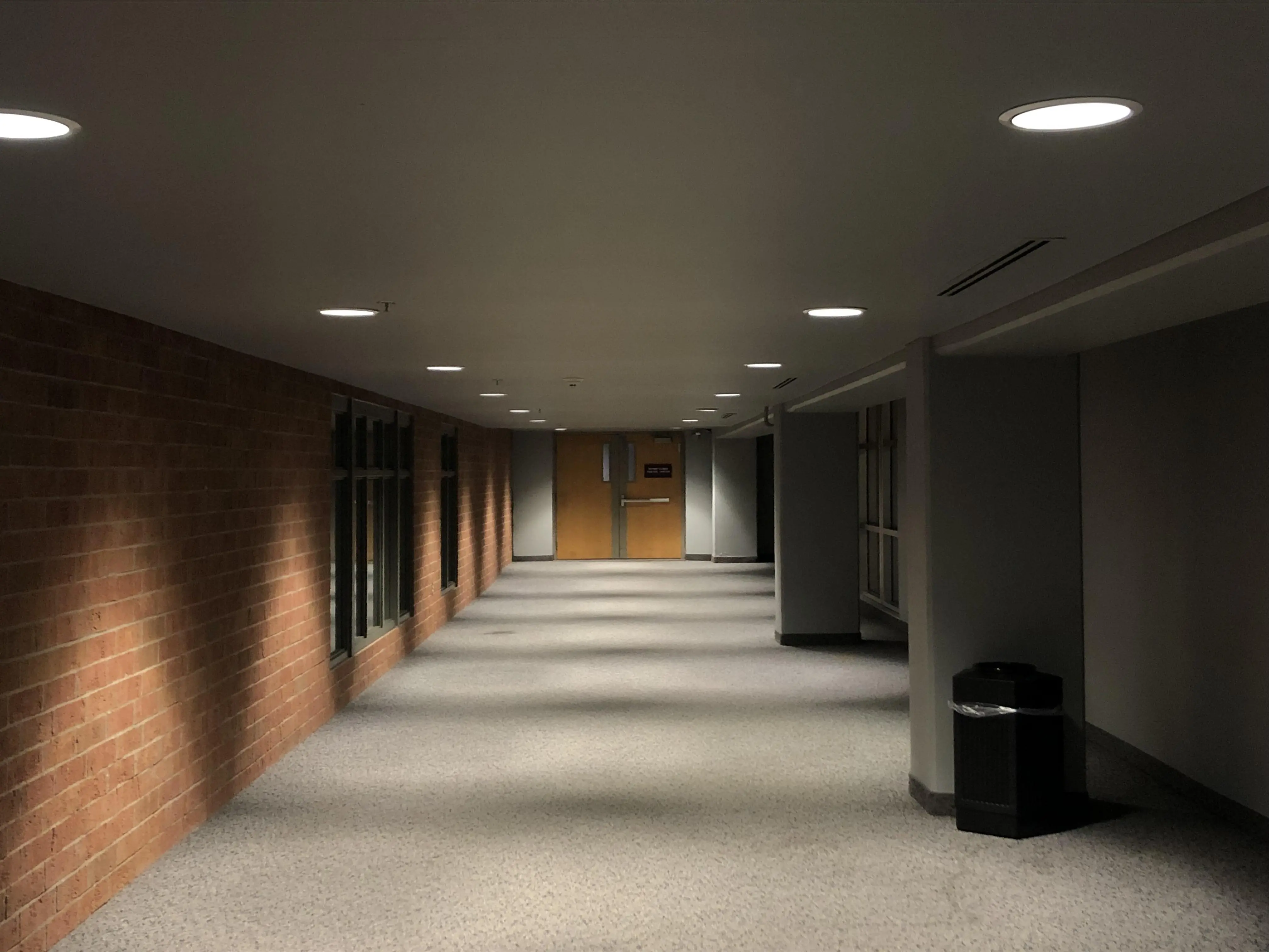 Empty interior corridor at night with a brick wall on the left, recessed ceiling lights, a speckled floor, and a single wooden door at the far end