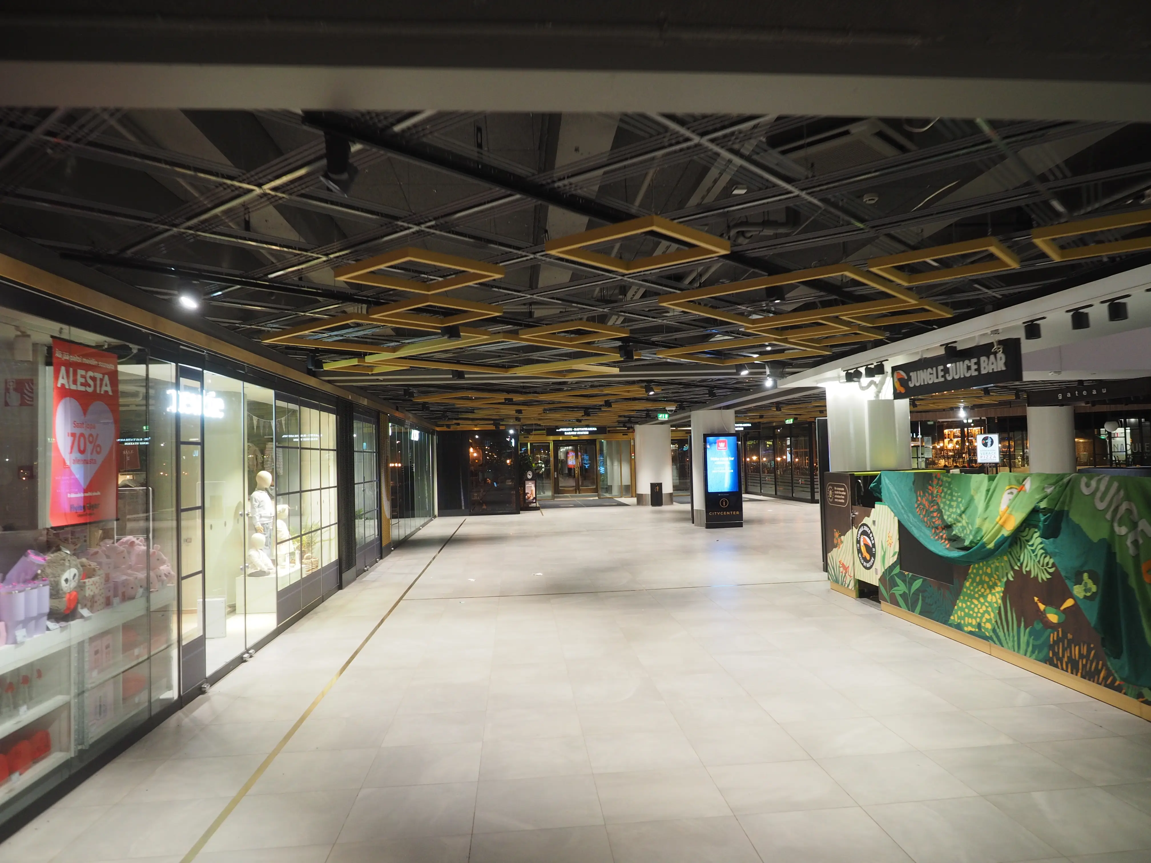 Empty shopping mall concourse at night, lit shop windows along one side, a juice bar along the other, and no people anywhere in the frame