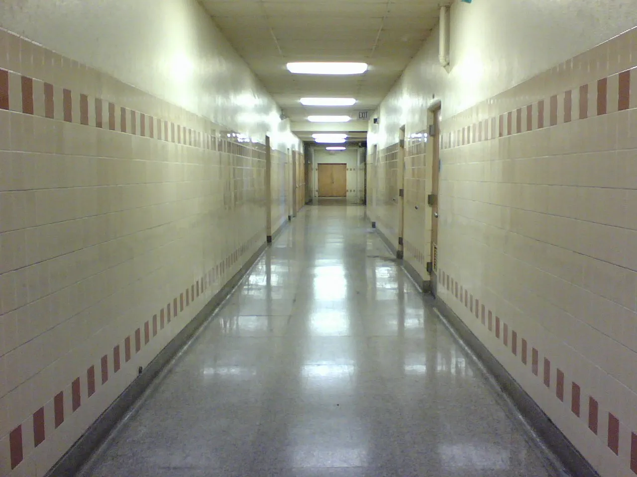 Empty school corridor with glazed tile walls, fluorescent ceiling lights, a polished terrazzo floor, and an exit sign at the far end
